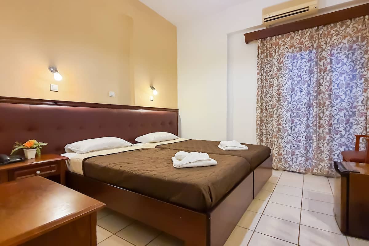 A hotel room with a large bed, a desk, and a chair. There is a window with curtains and an air conditioning unit above it.