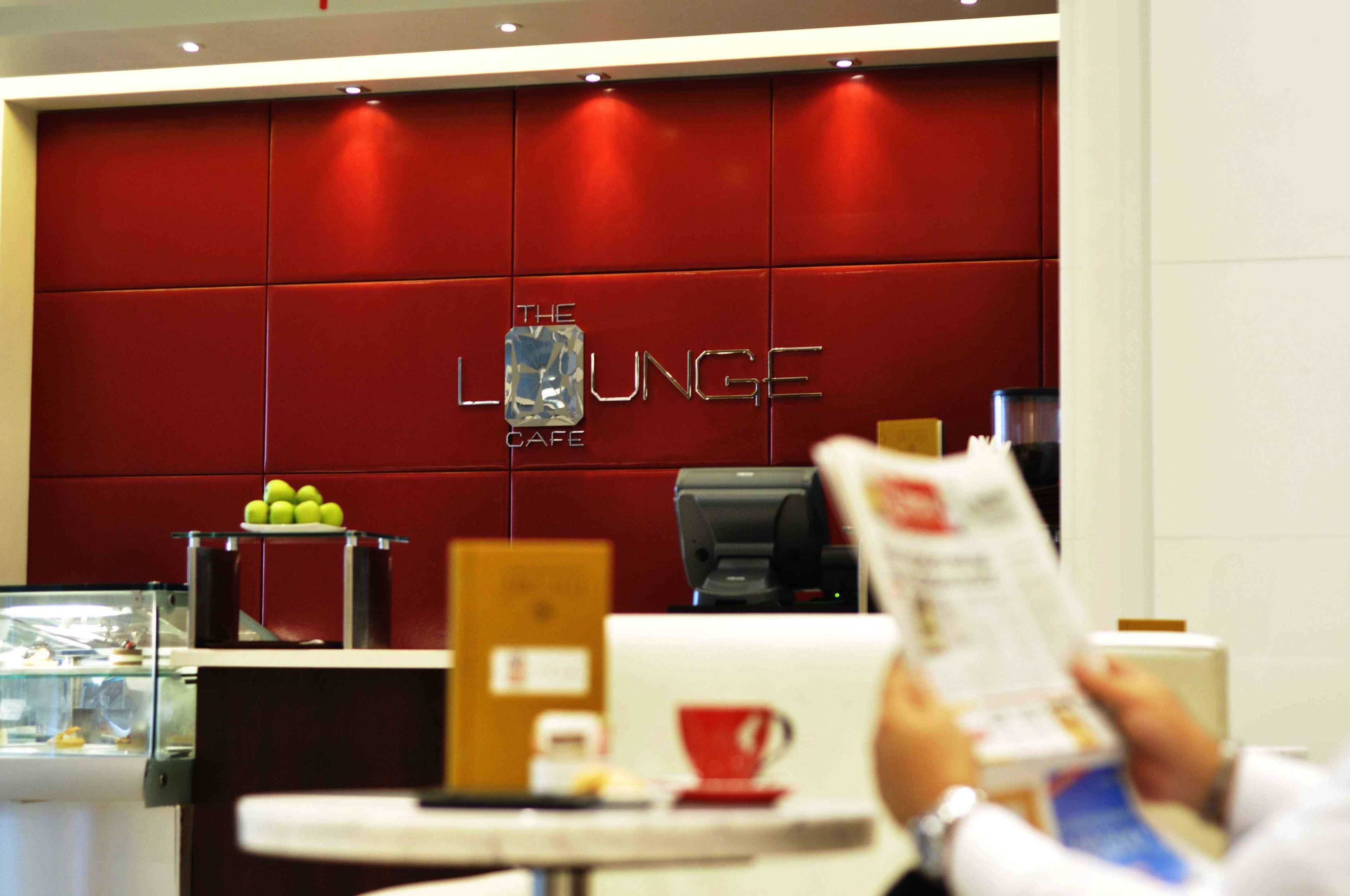 A red wall with "The Lounge Cafe" sign, a coffee machine, a menu stand, and a glass display case with apples.