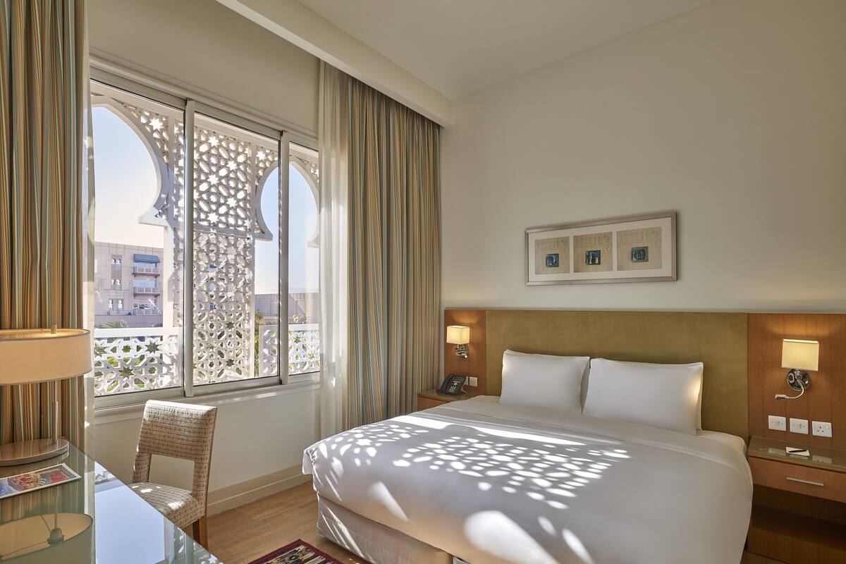 Habitación de hotel con una cama grande, un escritorio con lámpara, una silla, una mesita de noche con teléfono y una ventana con cortinas.