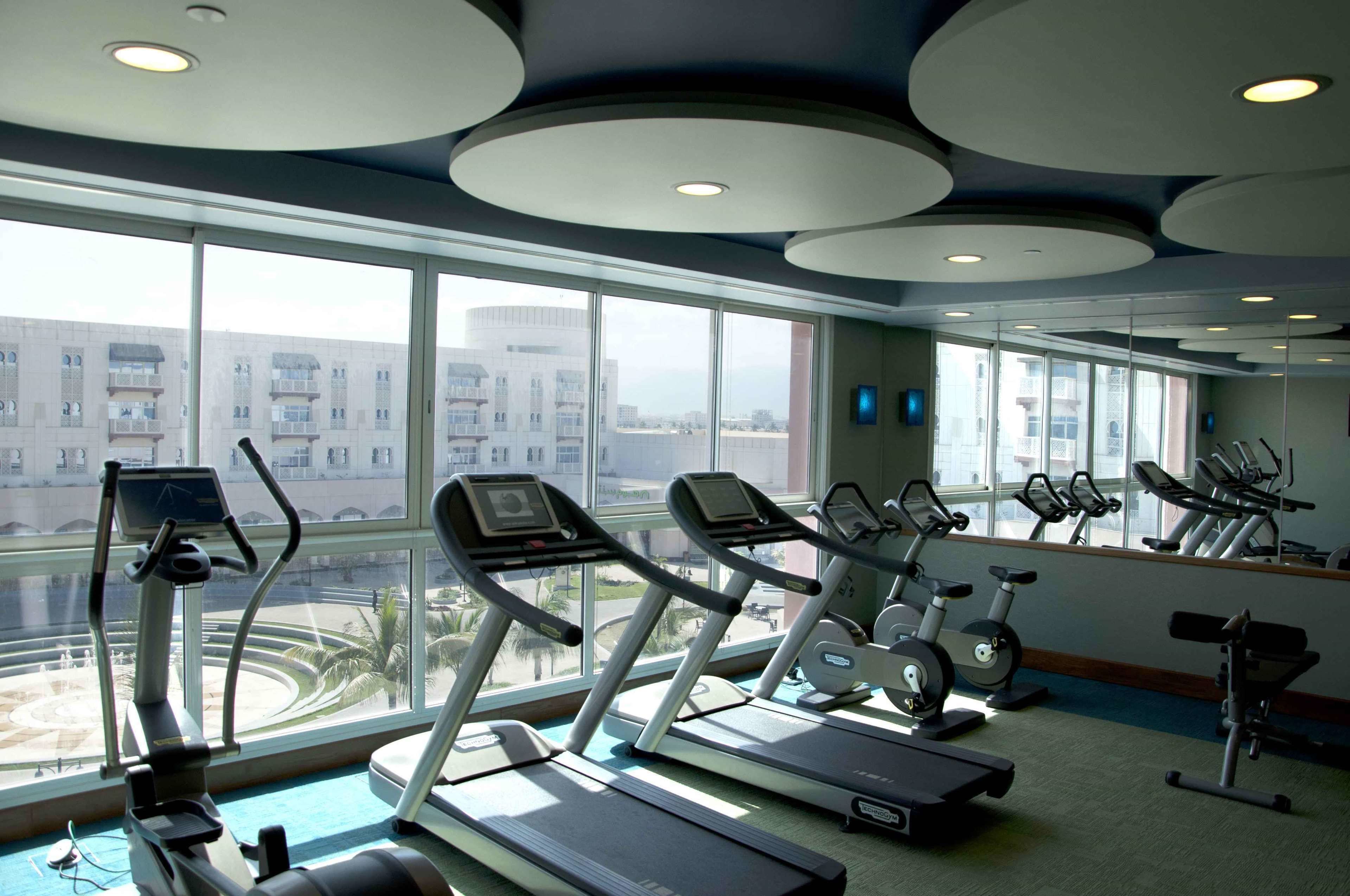 Gimnasio con cintas de correr, bicicletas estáticas y una máquina de remo. Cuenta con ventanas amplias que ofrecen vistas a edificios y palmeras.
