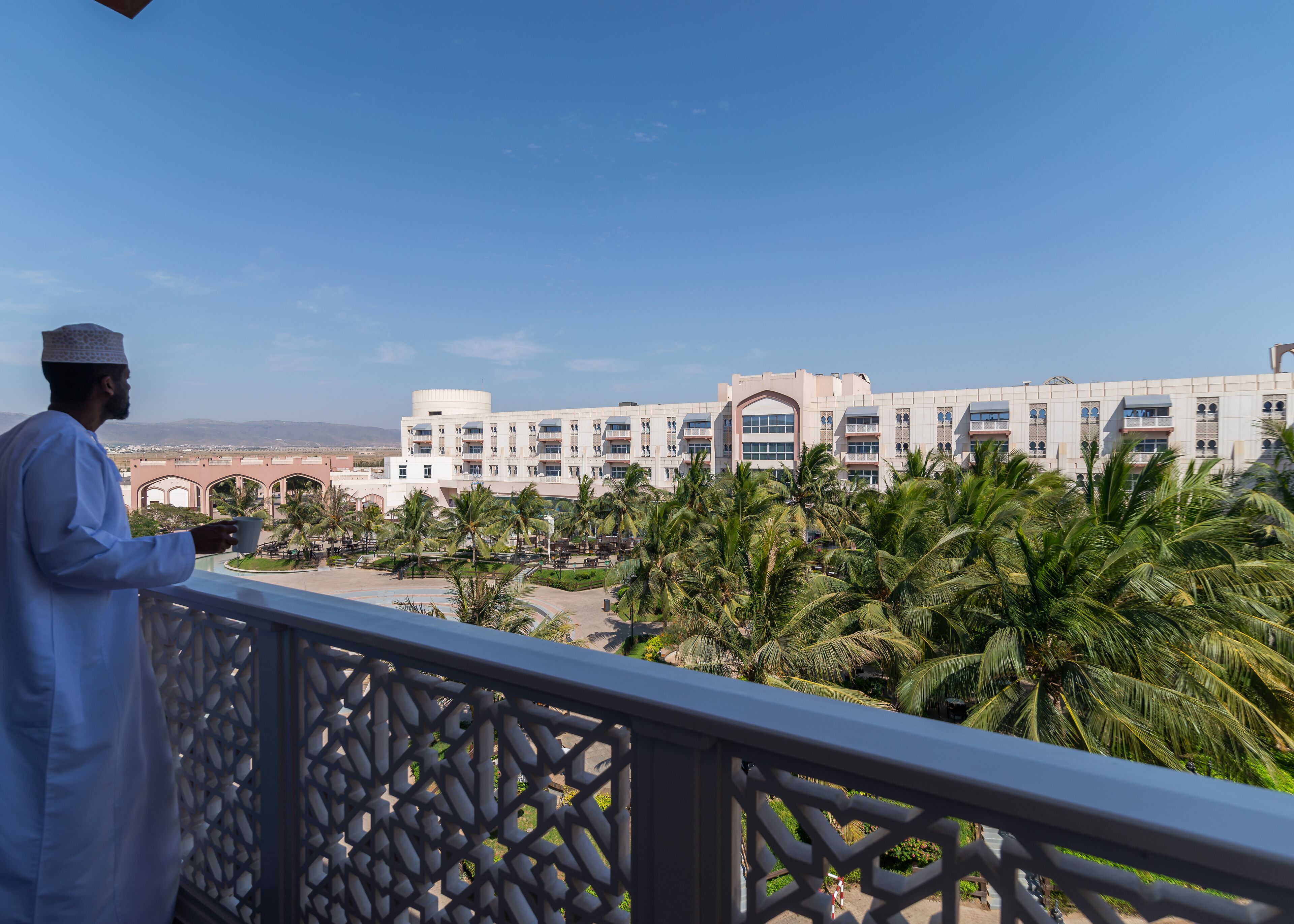 Un hombre en un balcón con vista a un hotel que tiene un patio, palmeras y montañas al fondo.