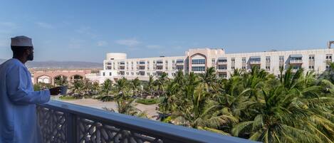 Un hombre en un balcón con vista a un hotel que tiene un patio, palmeras y montañas al fondo.