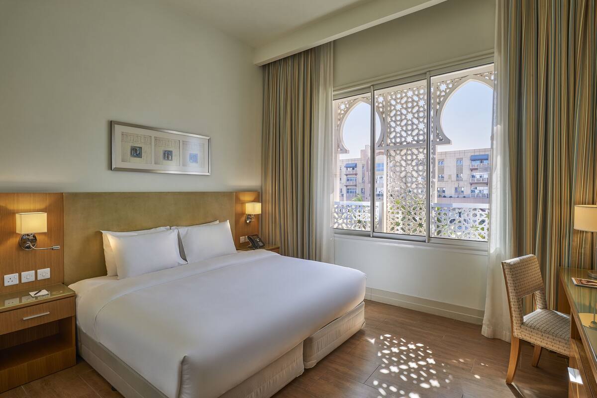 Habitación de hotel con una cama grande, dos lámparas de noche, una mesita de noche, una silla y una ventana con vistas a edificios.