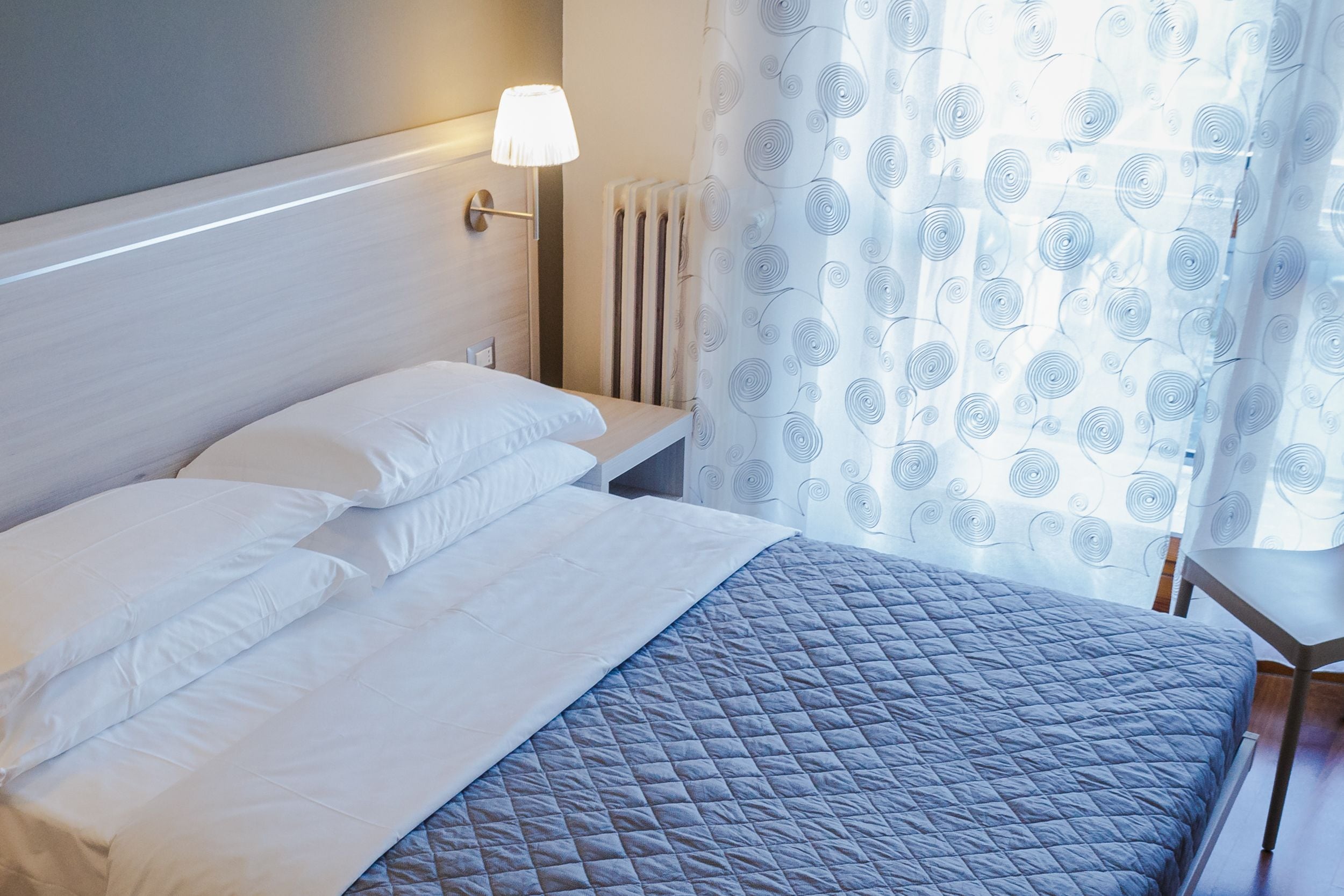 A neatly made bed with white and gray bedding, a bedside lamp, and a radiator.