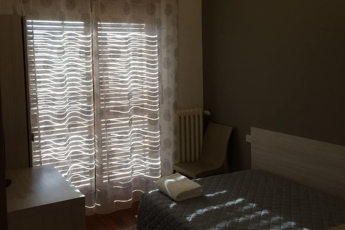 A bedroom with a bed, a chair, a radiator, and a window with patterned curtains.
