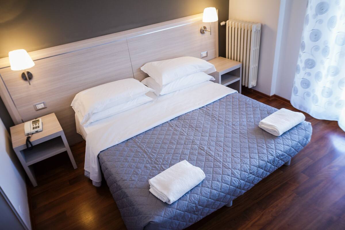 A neatly made bed with a quilted cover, two bedside tables with lamps, and a visible thermostat on the wall.