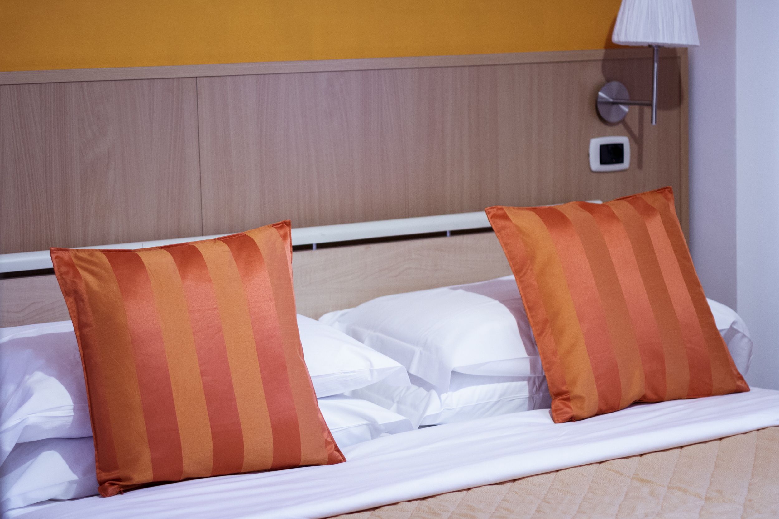 A hotel room with a bed, two striped pillows, a white headboard, a wall-mounted lamp, and a thermostat.