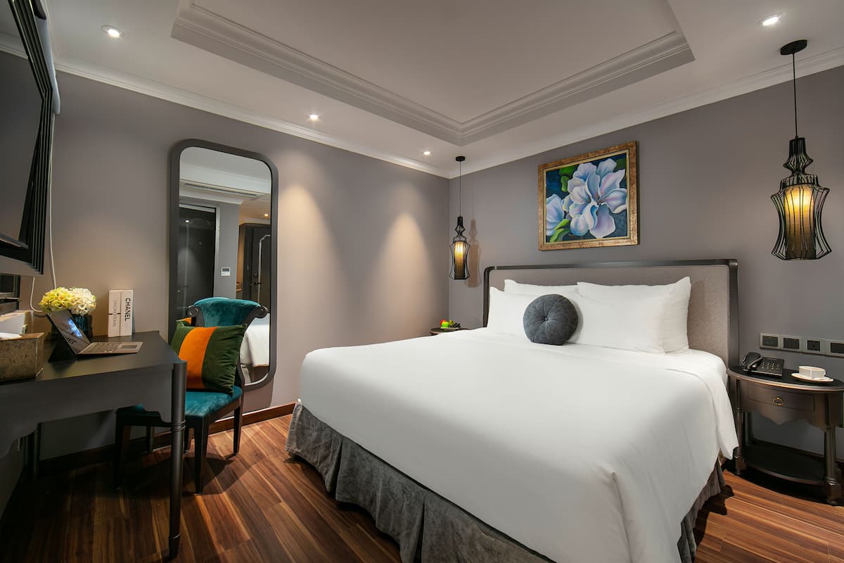 A modern hotel room with a large bed, a desk with a laptop, a chair, a bedside table with a phone, and a painting on the wall.
