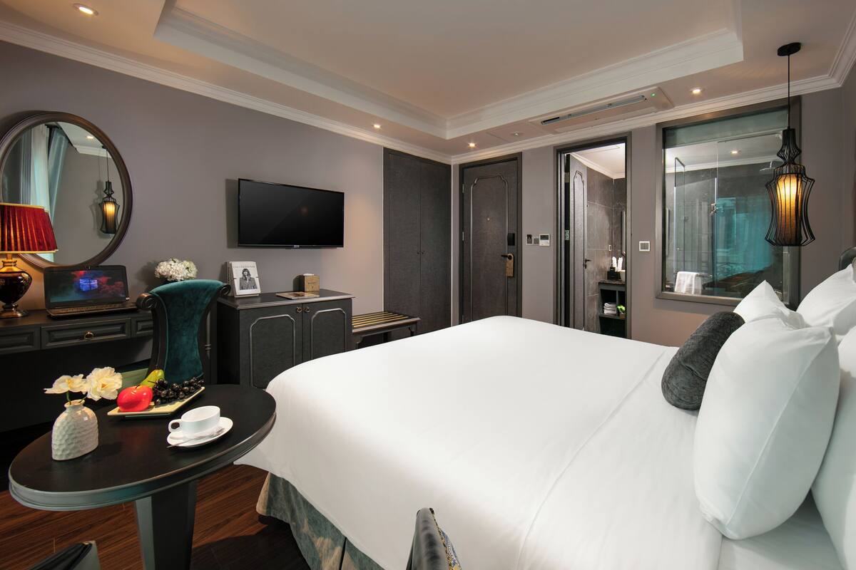 A hotel room with a large bed, a flat-screen TV, a desk with a lamp, and a small table with a vase and flowers.