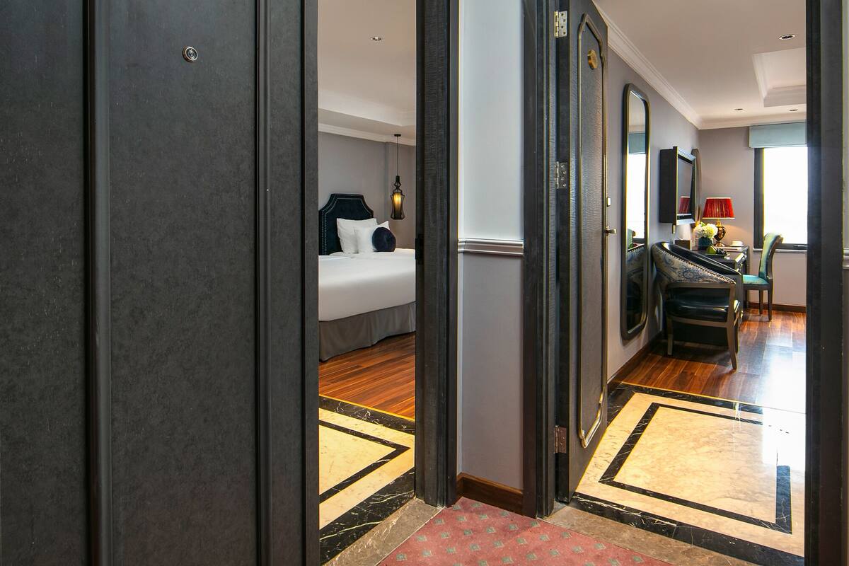 A hotel hallway with a red carpet, a bed with white linens, a chair, and a mirror on the wall.