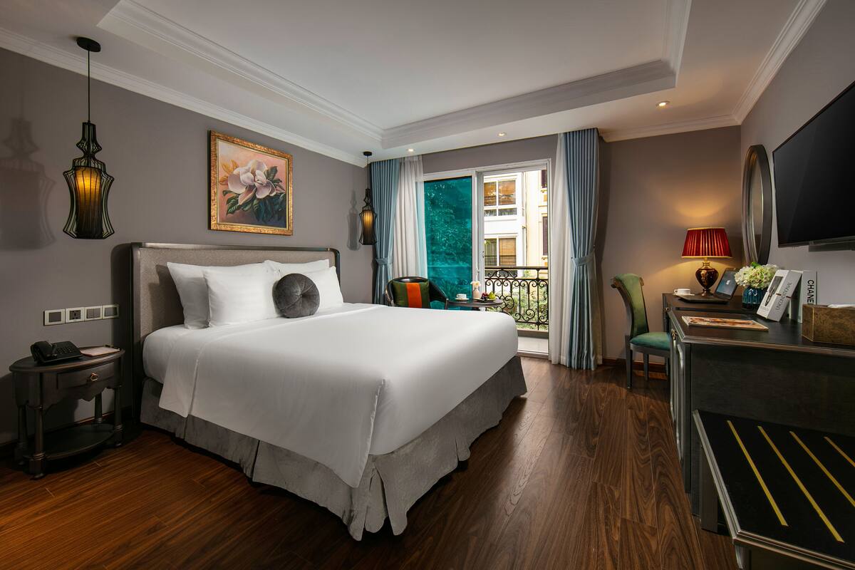 A hotel room with a large bed, a desk with a telephone, a television, and a balcony with a view of the city.