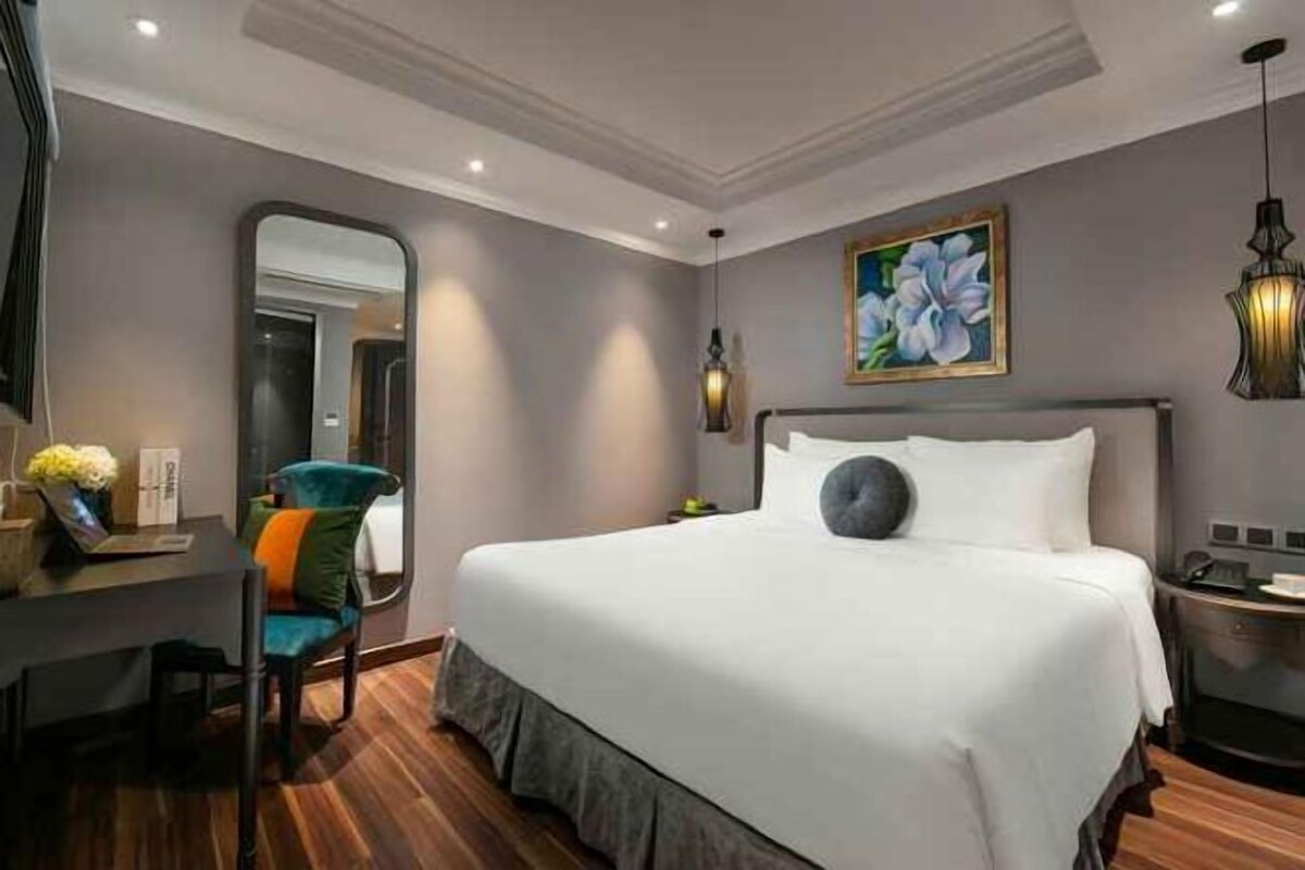 A hotel room with a large bed, a desk with a chair, a mirror, and a painting on the wall.
