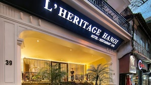 Entrada del hotel L'Heritage Hanoi con una imponente escalera, iluminación decorativa y un cartel que indica servicios de hotel, restaurante y spa.