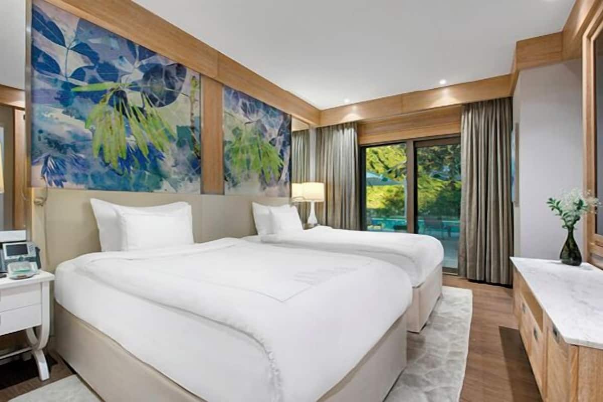 A hotel room with two beds, a large window, a painting, and a desk with a lamp.