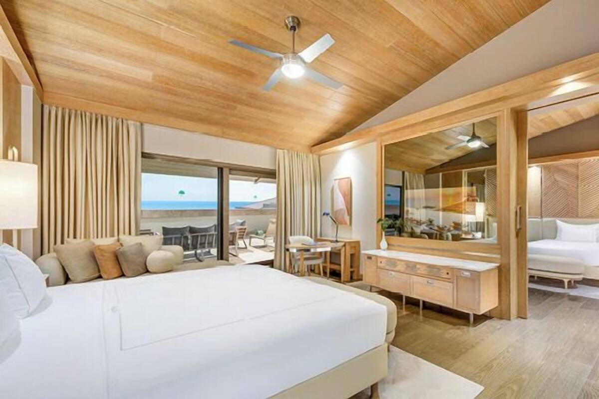 A hotel room with a large bed, a desk, a chair, a mirror, and a view of the beach.