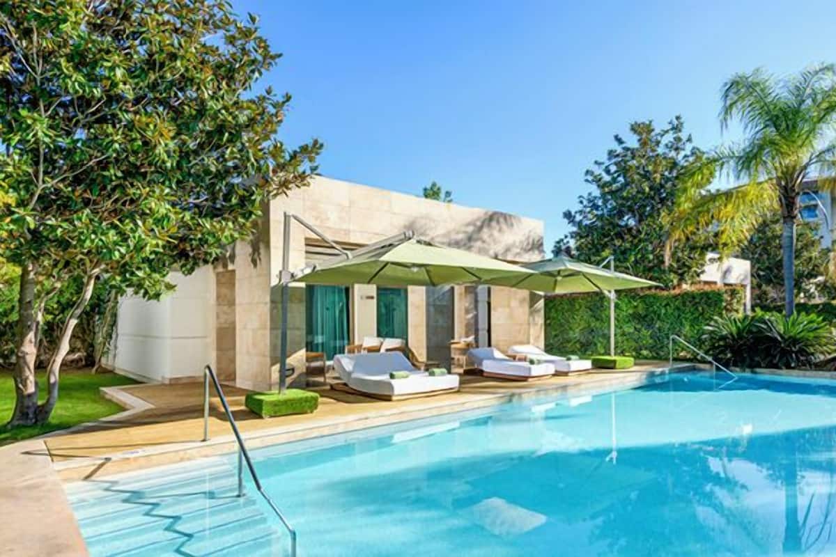 A swimming pool with lounge chairs and umbrellas, surrounded by greenery and a building.