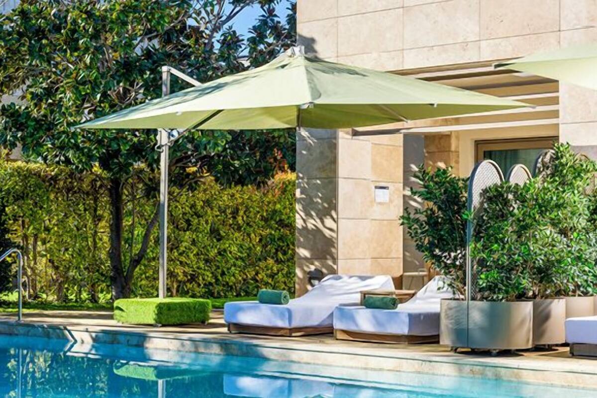 A swimming pool with lounge chairs and umbrellas, surrounded by greenery and a building.