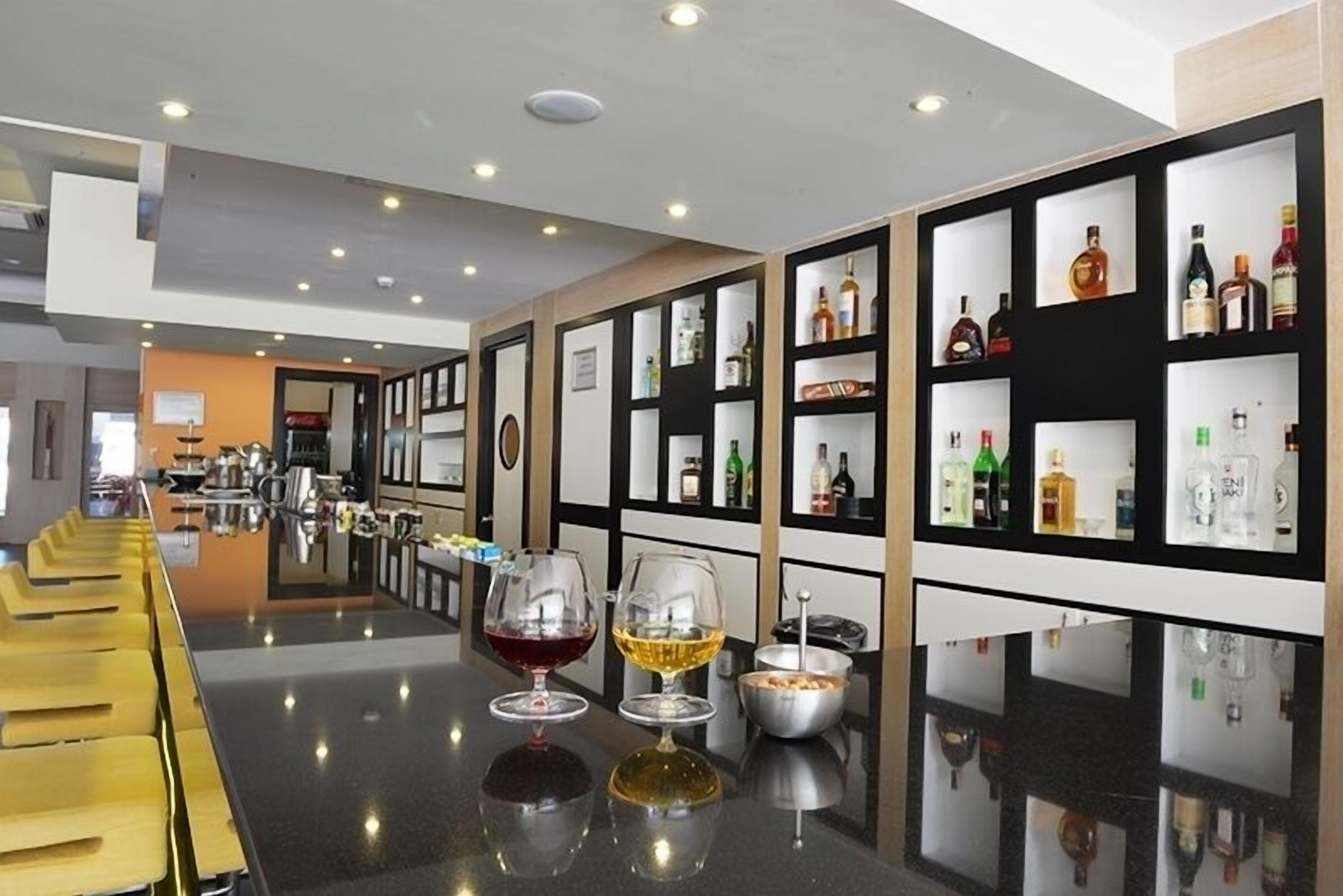 Una zona de bar con una barra alargada, varios estantes con botellas y dos copas de vino sobre la barra.