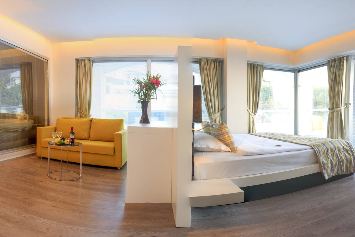 A modern bedroom with a sofa, a bedside table with fruit and wine, and a bed with a striped blanket.