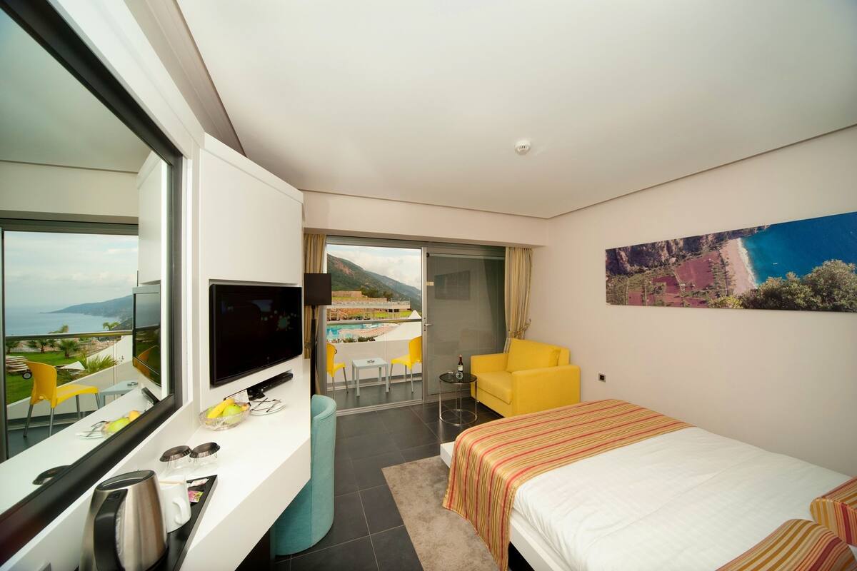 A modern hotel room with a large bed, a desk with a TV, a balcony with a view of the sea, and a painting on the wall.