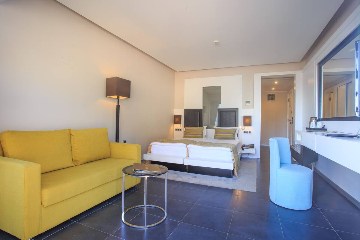 A modern hotel room with a yellow sofa, a round side table, a bed with white linens, a large mirror, and a blue chair.