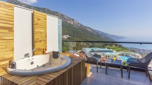 Uma varanda com hidromassagem, painéis de madeira e vista para uma vila costeira e montanhas.