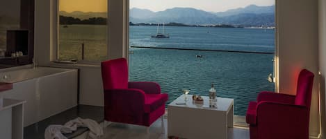 A modern room with a large window overlooking a body of water and mountains. There are two red chairs and a white coffee table.