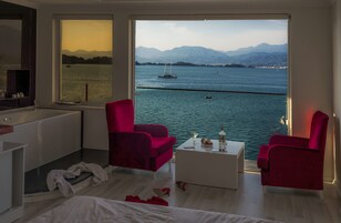 Una habitación moderna con un amplio ventanal que da a un cuerpo de agua y montañas. Hay dos sillas rojas y una mesa de centro blanca.