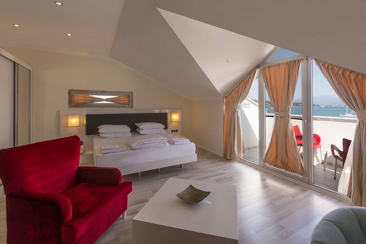 A hotel room with a bed, a red armchair, a white coffee table, and a view of the sea through large windows.