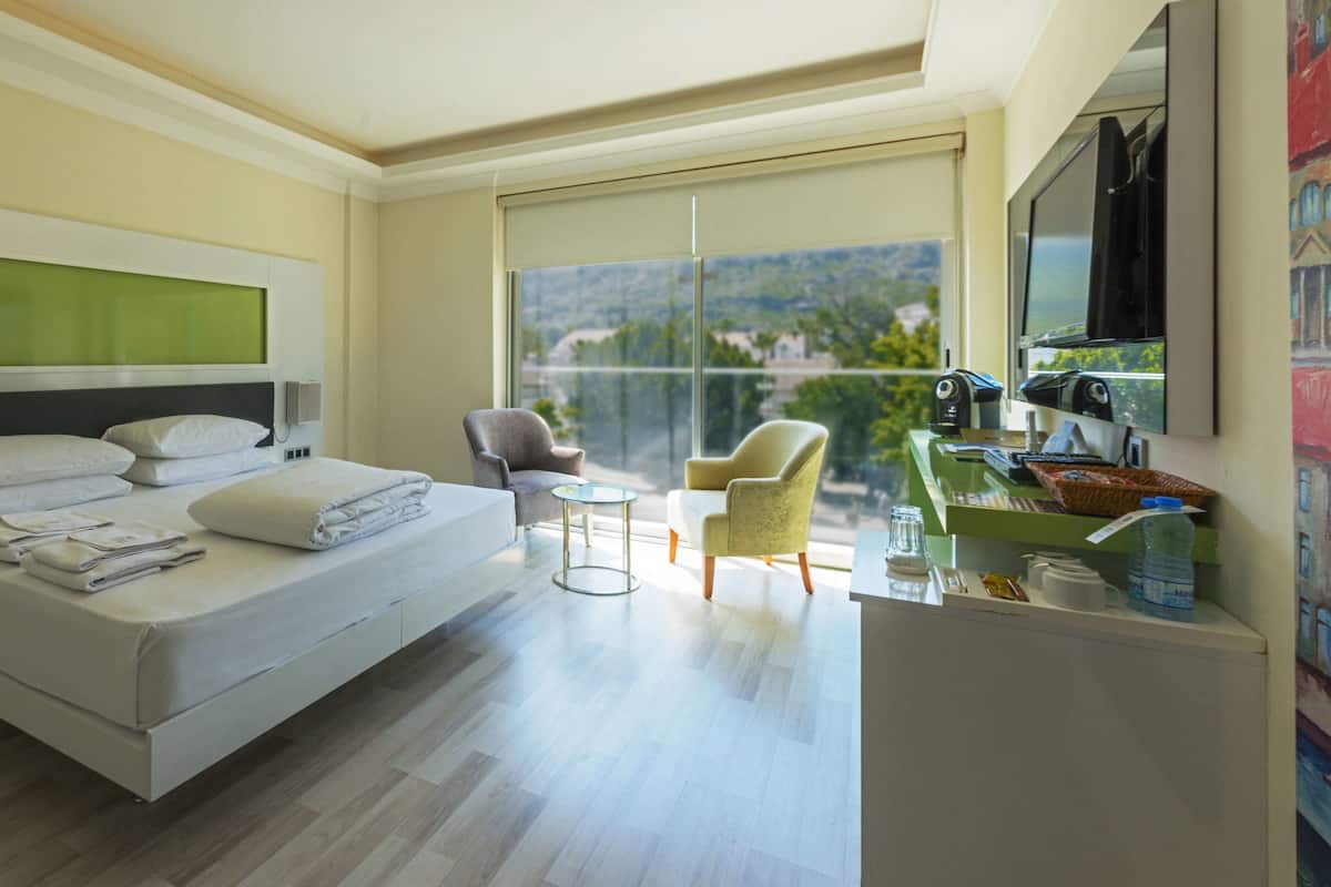 A modern hotel room with a large bed, a desk with a computer, a chair, and a view of the outdoors.
