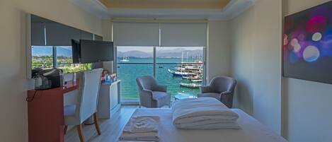 Habitación de hotel con cama, dos sillones, escritorio y televisor. Cuenta con un ventanal amplio con vistas a barcos y montañas.