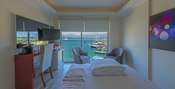 Habitación de hotel con cama, dos sillones, escritorio y televisor. Cuenta con un ventanal amplio con vistas a barcos y montañas.