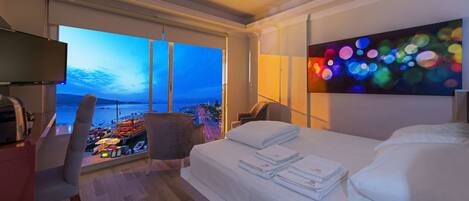 A hotel room with a bed, a chair, a desk, and a large window offering a view of a cityscape and water.