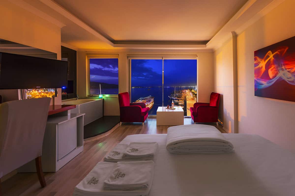 A hotel room with a bed, a desk, a chair, and a view of the marina at night.