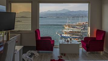 Een kamer met een groot raam met uitzicht op een jachthaven en bergen. Er staan twee rode stoelen, een witte salontafel en een televisie op een standaard.