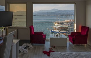 Een kamer met een groot raam met uitzicht op een jachthaven en bergen. Er staan twee rode stoelen, een witte salontafel en een televisie op een standaard.