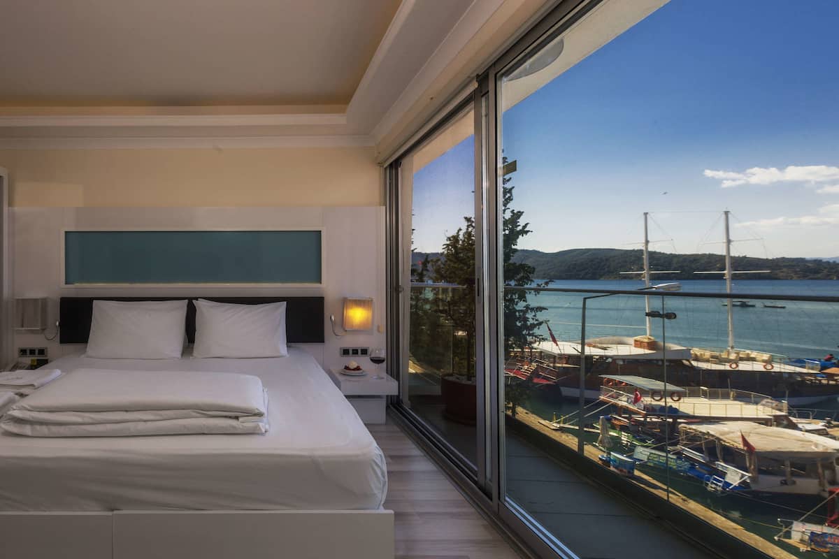 A modern hotel room with a large bed, a nightstand, a bedside table with a lamp, and a view of a marina with boats.