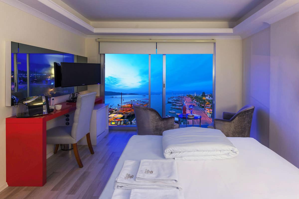 A hotel room with a bed, a desk with a TV, and a view of boats and a cityscape.