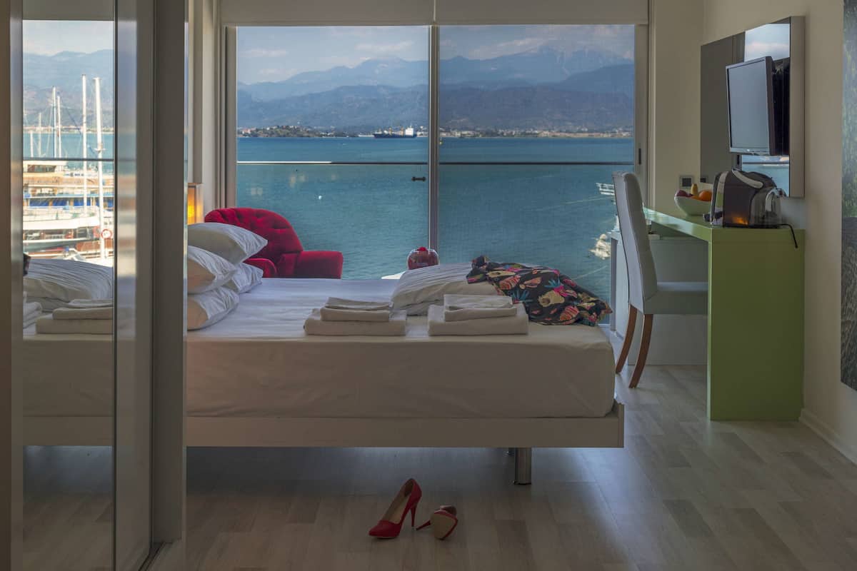 A hotel room with a bed, a desk, a chair, and a view of the sea and mountains.