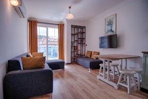 A modern living room with a sofa, a wooden dining table with stools, a bookshelf, and a TV mounted on the wall.