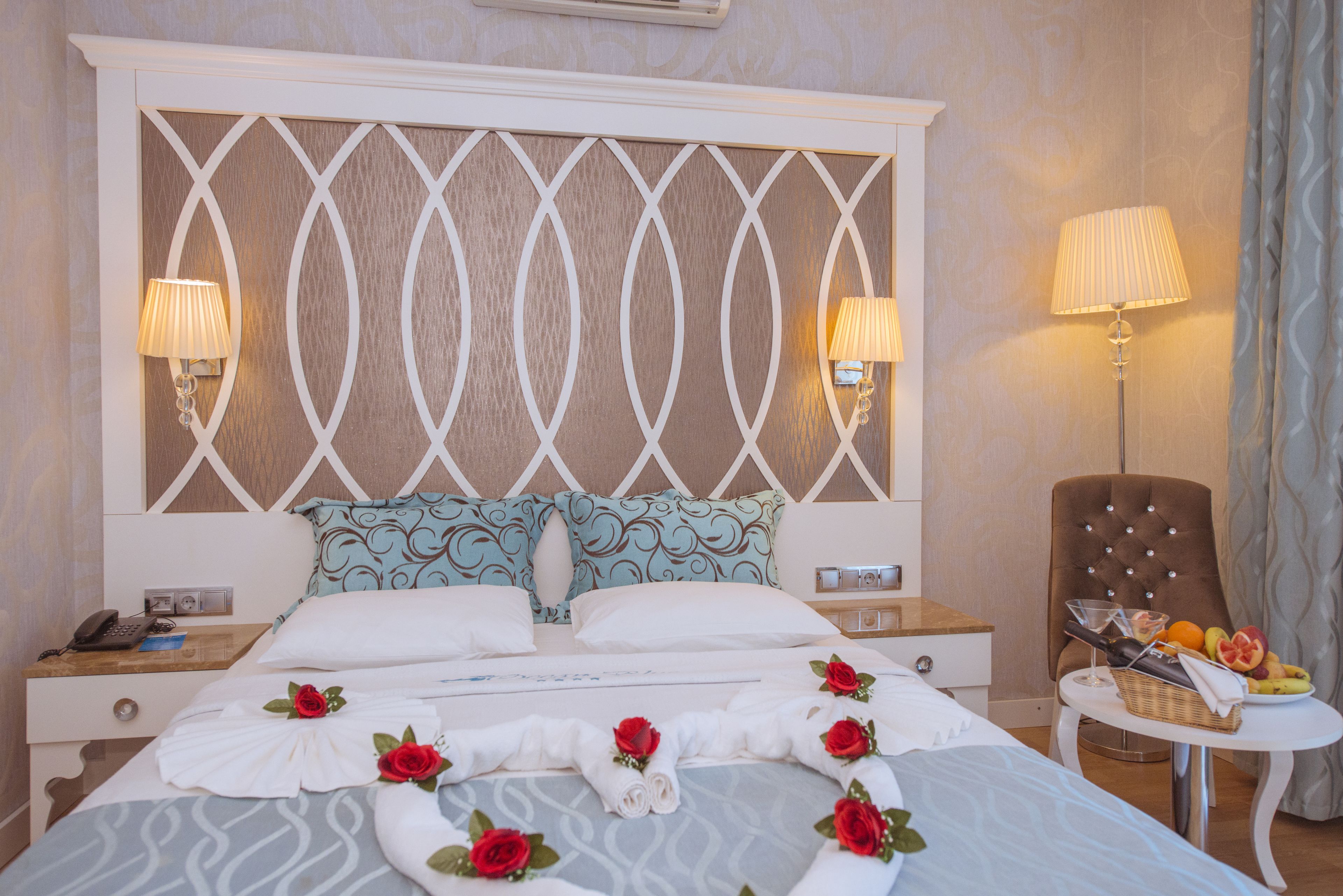 A hotel room with a large bed, two bedside tables, a nightstand, a chair, and a small table with a lamp and fruit.