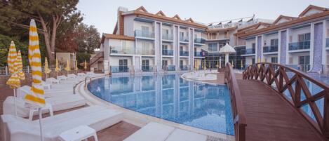 A swimming pool with lounge chairs and umbrellas, surrounded by a wooden deck and a building with multiple balconies.
