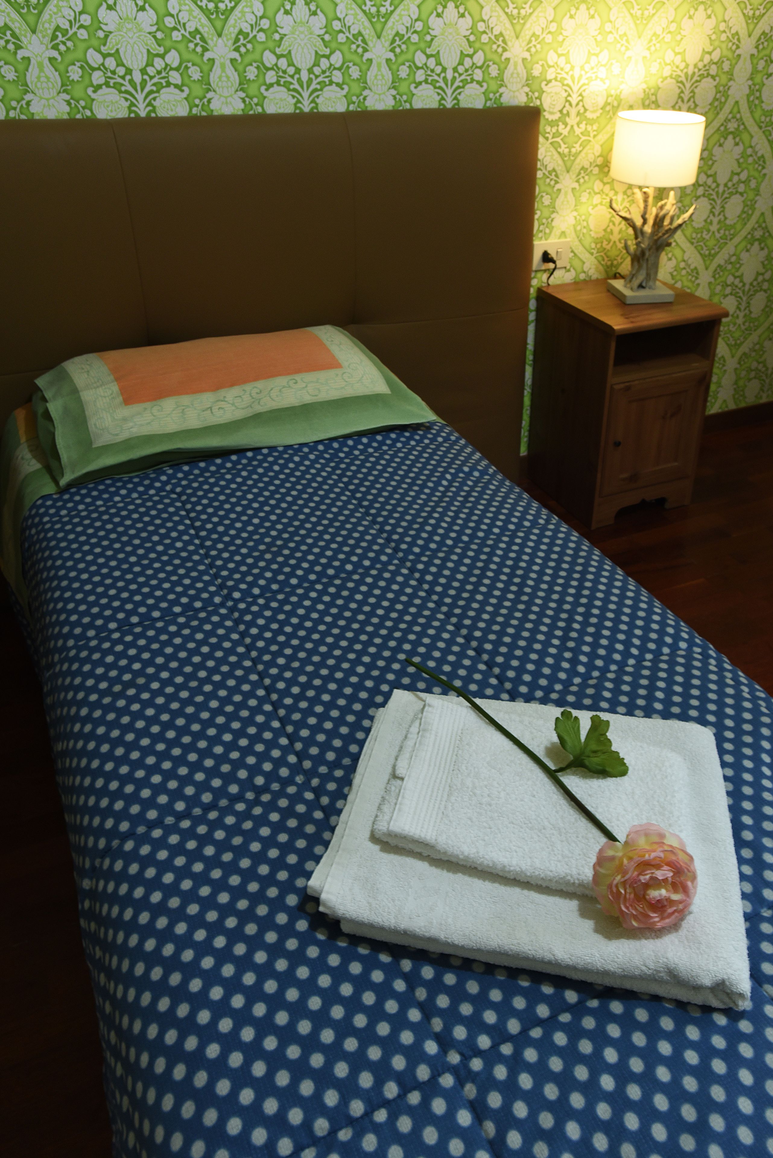 A neatly made bed with a blue polka dot bedspread, a white towel with a pink flower, and a wooden nightstand with a lamp.