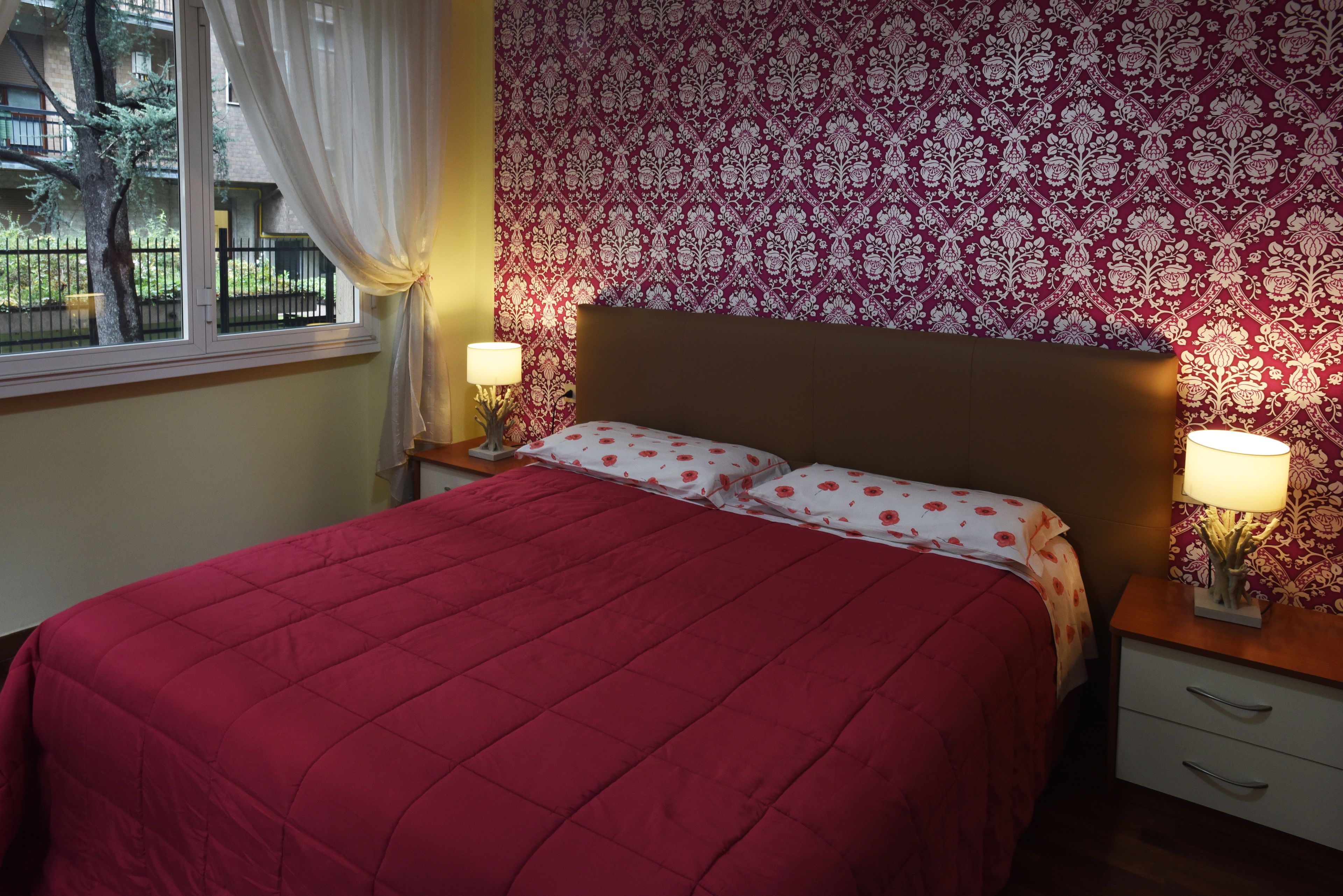 A bedroom with a large bed, two bedside tables with lamps, a patterned wallpaper, and a window with curtains.