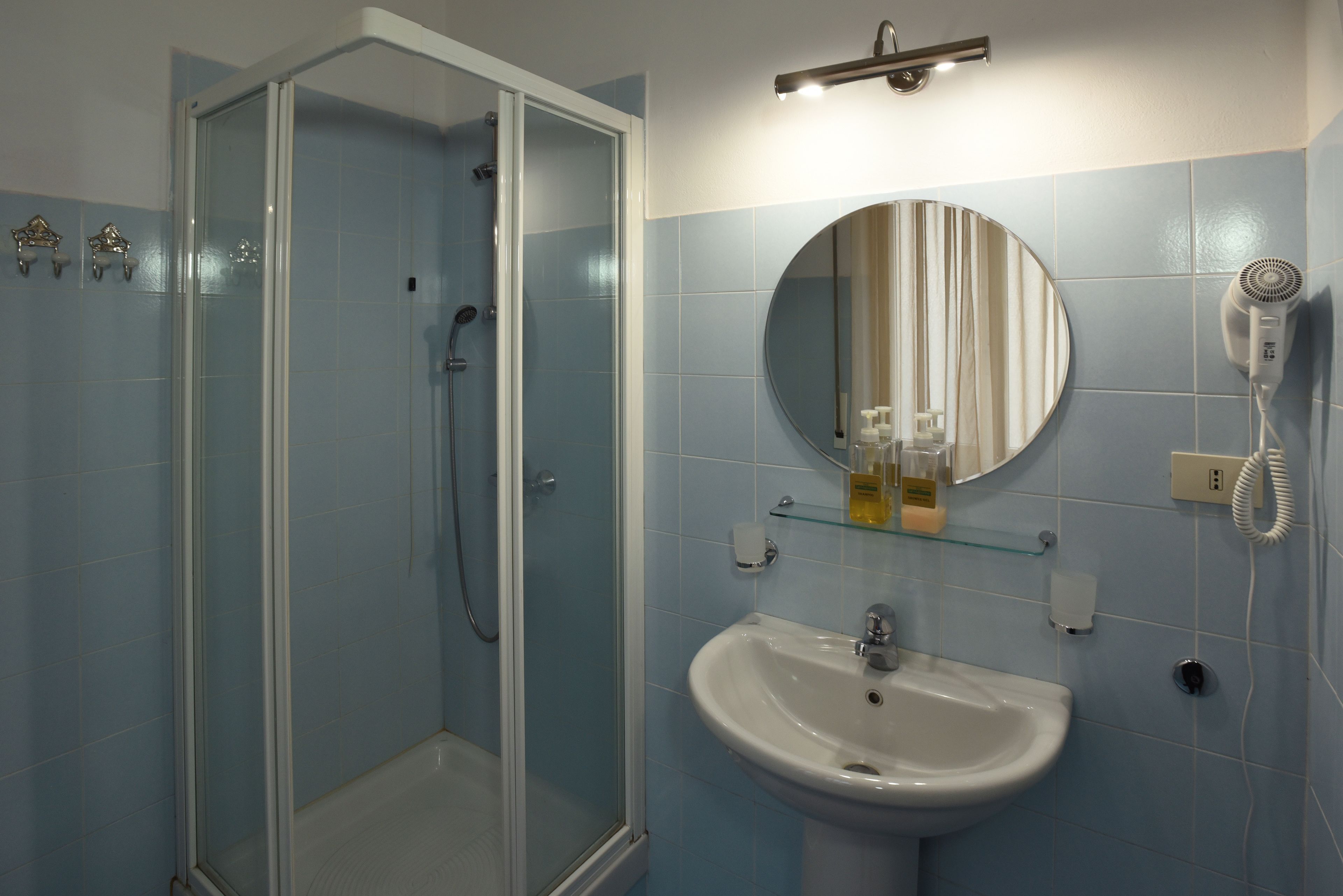 Een badkamer met een douche met glazen wand, een wit wastafelmeubel, een ronde spiegel en een haardroger aan de muur.