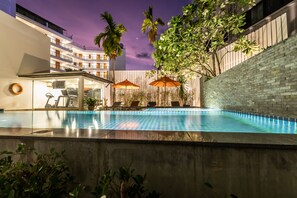 Una piscina al aire libre con sillones de descanso y sombrillas, rodeada por un edificio de gran altura con balcones y palmeras.
