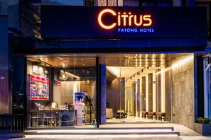 The exterior of the Citrus Patong Hotel with a modern design, featuring a glass entrance, a reception desk, and seating area visible through the glass doors.