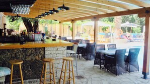 An outdoor patio area with a bar, stone flooring, wicker furniture, and a view of a pool and umbrellas. - Hotel Paloma Blanca (Corfu)