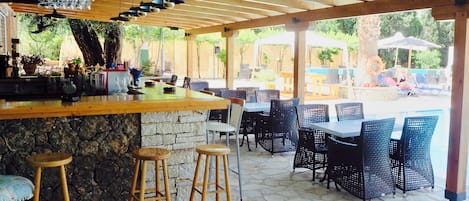 An outdoor patio area with a bar, stone flooring, wicker furniture, and a view of a pool and umbrellas.