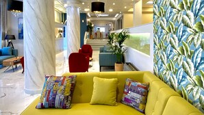 A modern hotel lobby with a yellow sectional sofa, marble columns, and a tropical leaf-patterned wall. - Hotel Paloma Blanca (Corfu)